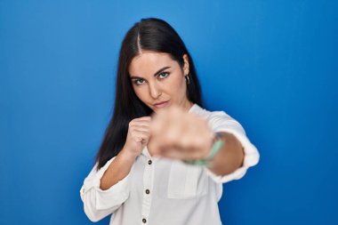 Young hispanic woman standing over blue background punching fist to fight, aggressive and angry attack, threat and violence 
