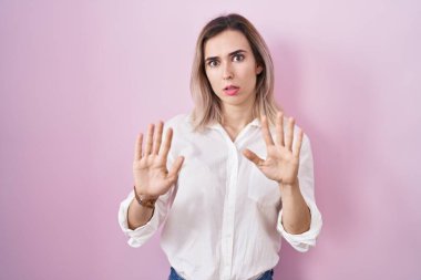 Young beautiful woman standing over pink background moving away hands palms showing refusal and denial with afraid and disgusting expression. stop and forbidden. 