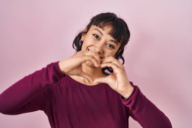 Pembe arka planda duran genç ve güzel bir kadın. Sevgiyle gülümsüyor. Elleriyle kalp sembolü yapıyor. romantik kavram. 