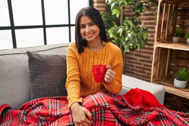 Genç İspanyol kadın kanepede oturuyor ve yüzünde mutlu ve havalı bir gülümsemeyle evde kahve içiyor. şanslı kişi. 