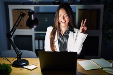 Geceleri ofiste çalışan genç esmer kadın dizüstü bilgisayarla gülümseyen ve kameraya göz kırpan parmaklarıyla zafer işareti yapan. İki numara.. 