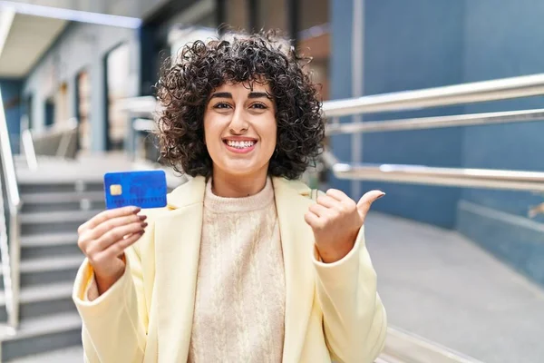 Young brunette woman with curly hair holding credit card pointing thumb up to the side smiling happy with open mouth