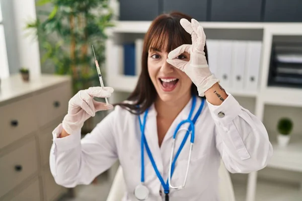 Young brunette doctor woman holding syringe smiling happy doing ok sign with hand on eye looking through fingers