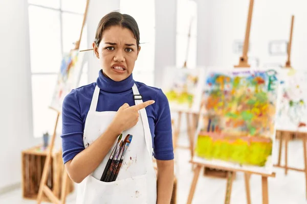 Young brunette woman at art studio pointing aside worried and nervous with forefinger, concerned and surprised expression