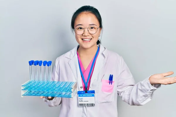 Young chinese girl wearing scientist uniform holding test tube celebrating achievement with happy smile and winner expression with raised hand