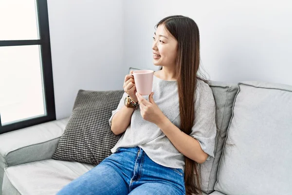 Genç Asyalı kadın oturma odasındaki kanepede kahve içiyor.