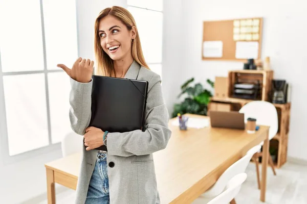 Blonde business woman at the office smiling with happy face looking and pointing to the side with thumb up.