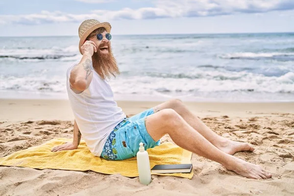 Genç kızıl saçlı adam kumsalda havlunun üzerinde oturmuş akıllı telefondan konuşuyor..