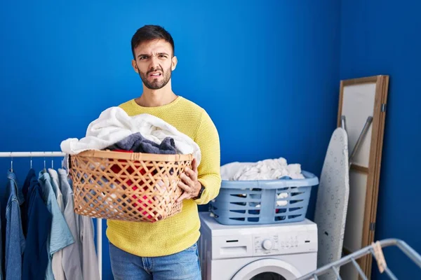 Hispanic man holding laundry basket clueless and confused expression. doubt concept.
