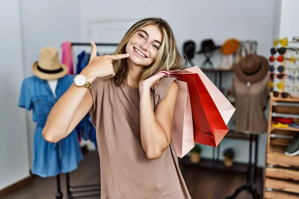 年轻的金发女人拿着购物袋在零售店里欢快地微笑着 用手指 牙齿和嘴指指点着 牙齿健康概念 — 图库照片