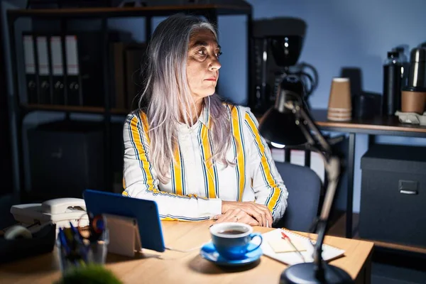 Middle age woman with grey hair working at the office at night looking to side, relax profile pose with natural face with confident smile.
