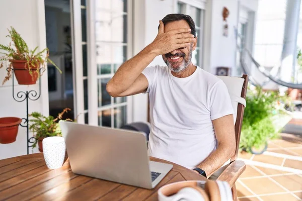 Middle age man using computer laptop at home smiling and laughing with hand on face covering eyes for surprise. blind concept.