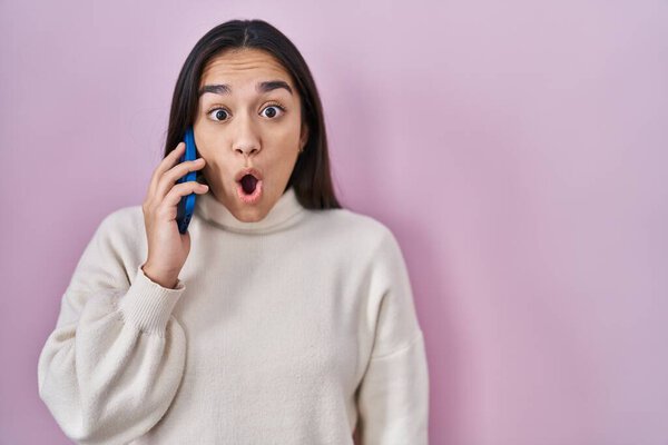 Young south asian woman having conversation talking on the smartphone scared and amazed with open mouth for surprise, disbelief face 