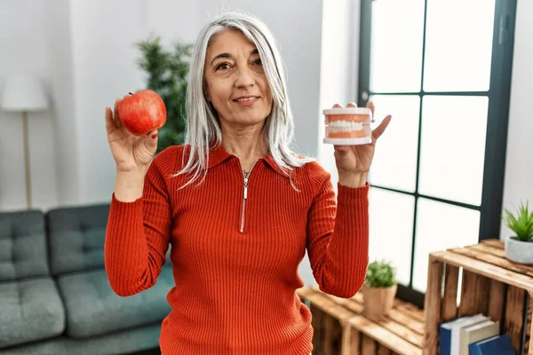 Middle age grey-haired woman smiling confident holding red apple and denture at home