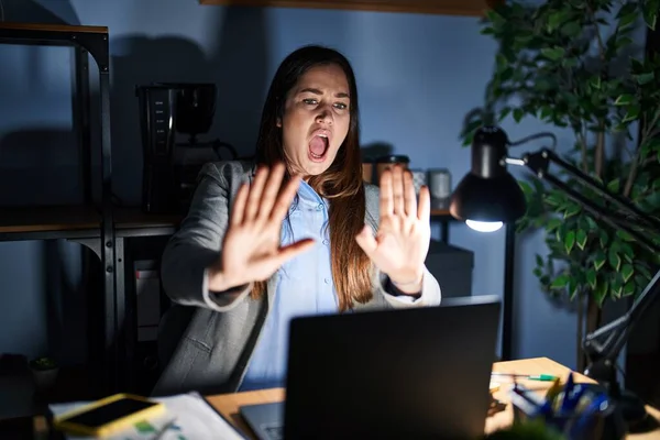 Young brunette woman working at the office at night afraid and terrified with fear expression stop gesture with hands, shouting in shock. panic concept.