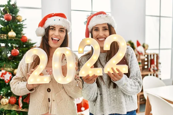 Two women celebrate new year holding 2022 lights standing by christmas tree at home
