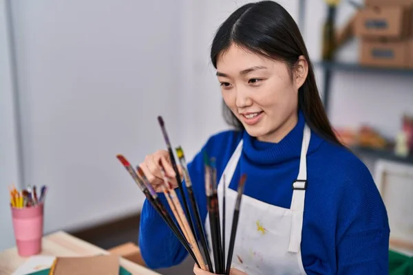 Chinese woman artist holding paintbrushes sitting on table at art studio