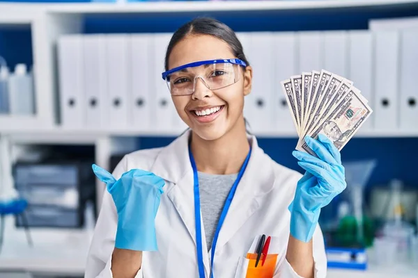 Young brazilian woman working at scientist laboratory holding money pointing thumb up to the side smiling happy with open mouth