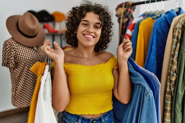 Young latin customer woman smiling happy holding hanger with clothes at clothing store.