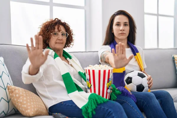 Hispanic mother and daughter watching football supporting team with open hand doing stop sign with serious and confident expression, defense gesture