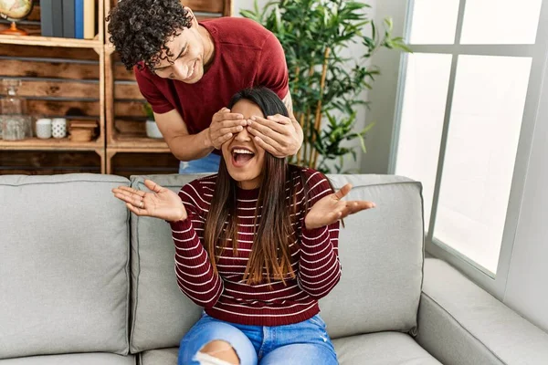 Man surprising his girlfriend with hands on eyes sitting on the sofa at home.