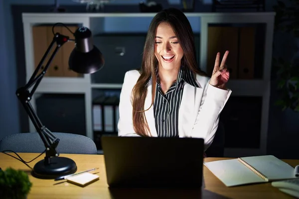 Young brunette woman working at the office at night with laptop smiling with happy face winking at the camera doing victory sign with fingers. number two.
