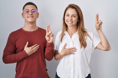 Mother and son standing together over isolated background smiling swearing with hand on chest and fingers up, making a loyalty promise oath 