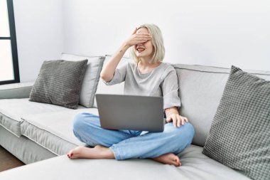 Young caucasian woman using laptop at home sitting on the sofa smiling and laughing with hand on face covering eyes for surprise. blind concept. 