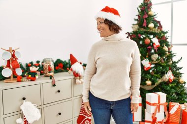 Orta yaşlı İspanyol kadın noel ağacının yanında duruyor. Yüzünde gülümseme ve doğal bir ifadeyle yan yana bakıyor. Kendinden emin gülen. 