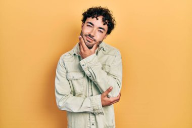 Gündelik kıyafetler giyen genç İspanyol adam kameraya kendinden emin bakıyor kollarını kavuşturup gülümsüyor ve çenesini kaldırıyor. Olumlu düşünmek. 