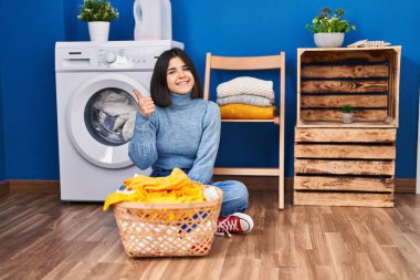 Young hispanic woman at laundry room smiling happy and positive, thumb up doing excellent and approval sign 