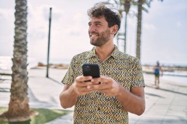 Genç İspanyol adam deniz kenarında akıllı telefon kullanarak gülümsüyor.