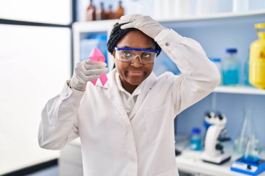 African american woman working at scientist laboratory holding pink ribbon stressed and frustrated with hand on head, surprised and angry face 