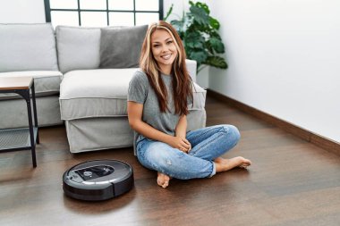 Young caucasian woman sitting at home by vacuum robot looking positive and happy standing and smiling with a confident smile showing teeth 