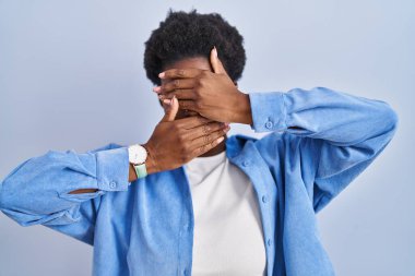 Mavi arka planda dikilen Afro-Amerikalı kadın gözlerini ve ağzını elleriyle kapatıyor, şaşırmış ve şok olmuş. duygu gizleme 