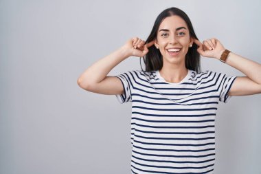 Young brunette woman wearing striped t shirt smiling pulling ears with fingers, funny gesture. audition problem 