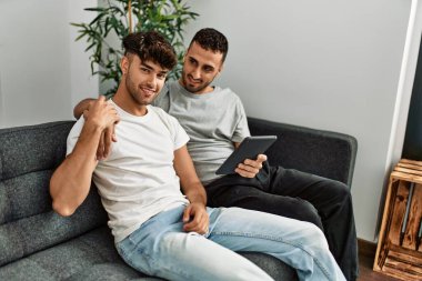 Two hispanic men couple using touchpad hugging each other at home