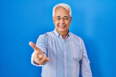 Hispanic senior man wearing glasses smiling cheerful offering palm hand giving assistance and acceptance. 
