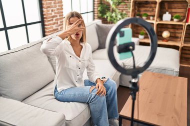 Young blonde woman recording vlog tutorial with smartphone at home peeking in shock covering face and eyes with hand, looking through fingers afraid 
