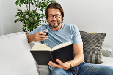 Orta yaşlı beyaz bir adam kitap okuyor evde şarap içiyor.