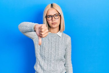 Beautiful blonde woman wearing glasses looking unhappy and angry showing rejection and negative with thumbs down gesture. bad expression. 