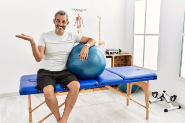 Orta yaşlı İspanyol adam ağrı kesici kliniğinde elinde pilates topu tutarken avuç içi açık bir şekilde fotokopi alanını gösteriyor. 