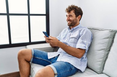 Genç İspanyol adam akıllı telefon kullanıyor. Evdeki kanepede oturuyor..