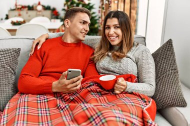 Akıllı telefon kullanan ve kahve içen genç bir çift. Evdeki kanepede oturmak..