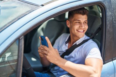 Young hispanic man using smartphone sitting on car at street