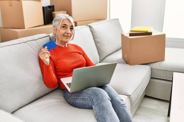 Middle age grey-haired woman smiling confident using laptop and credit card at new home