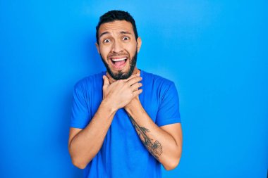 Hispanic man with beard wearing casual blue t shirt shouting and suffocate because painful strangle. health problem. asphyxiate and suicide concept. 