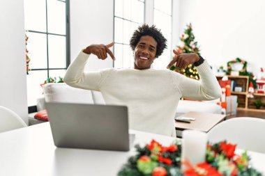Genç Afro-Amerikalı adam dizüstü bilgisayar kullanıyor. Noel ağacının yanında oturuyor. Yüzünde gülümsemeyle kendinden emin görünüyor.. 