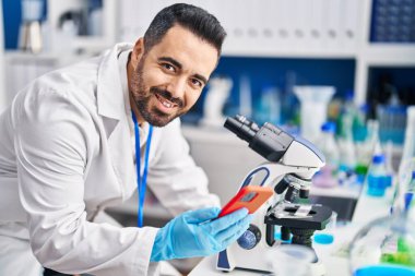 Young hispanic man scientist using microscope and smartphone at laboratory