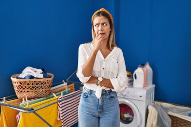 Young blonde woman at laundry room looking stressed and nervous with hands on mouth biting nails. anxiety problem. 